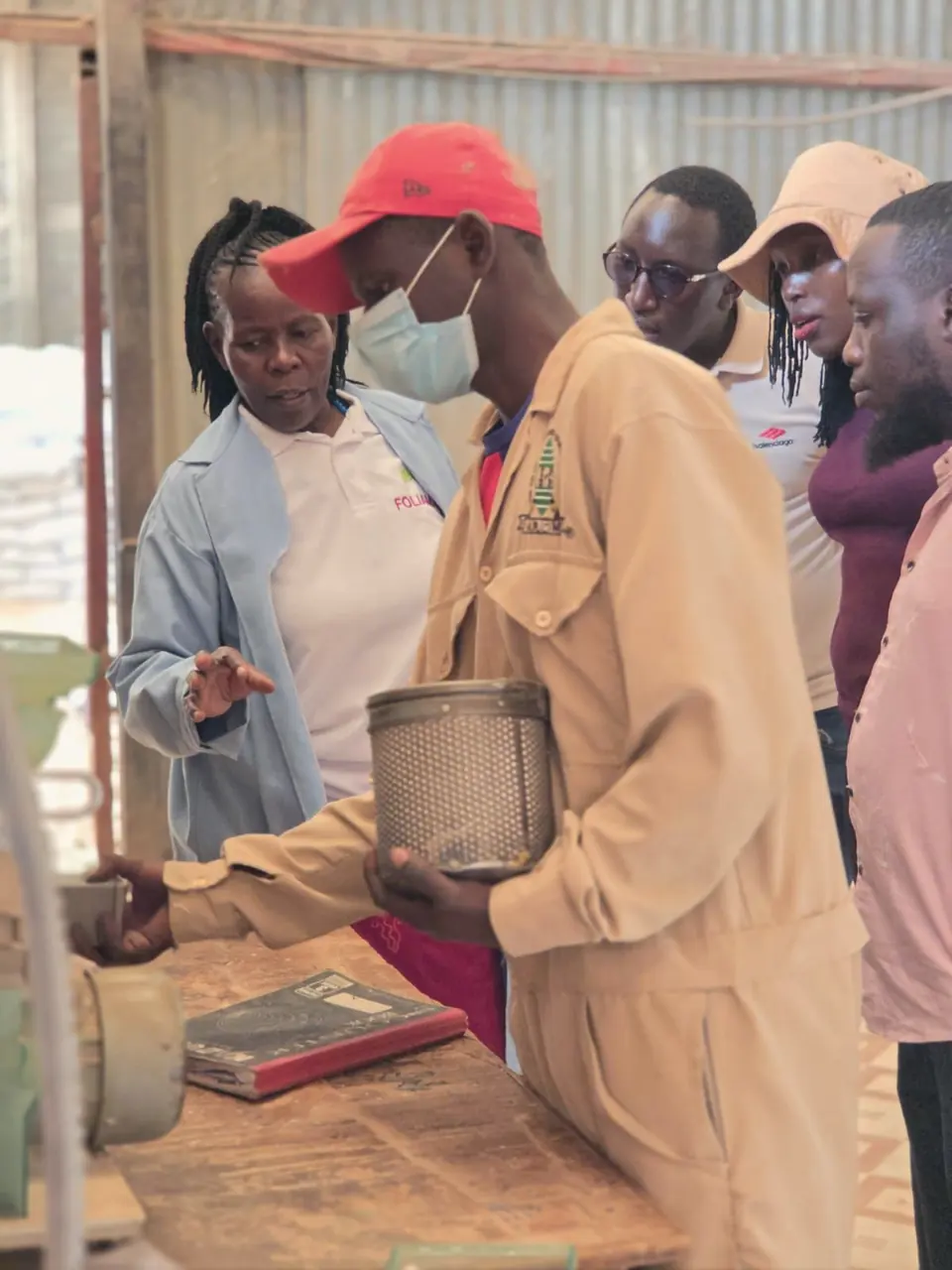 A partner technician holds a mesh sieve at a workbench while the ENKOP team and partner staff look on.